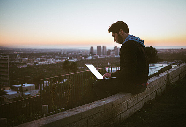 a man sitting on the edge of a wall outside on his laptop